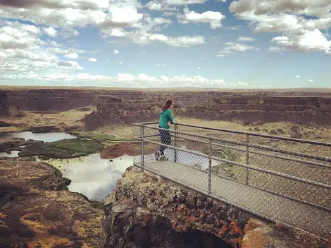 A person stands on a fenced-in overlook viewing a vast landscape with lakes and cliffs under a cloudy sky.