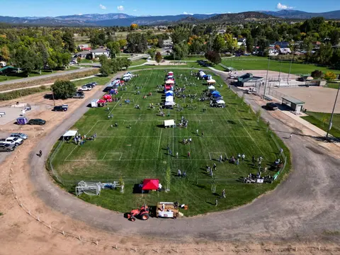 An aerial view shows an outdoor community event with tents and people gathered on a large grassy field.