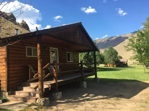 A wooden cabin sits in a grassy valley with mountains in the background under a blue sky.