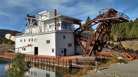 A large historic gold dredge sits on the water next to a wooden walkway.