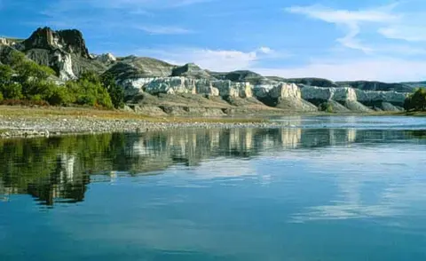 A wide river with calm water reflects white chalk cliffs and green hills under a blue sky.