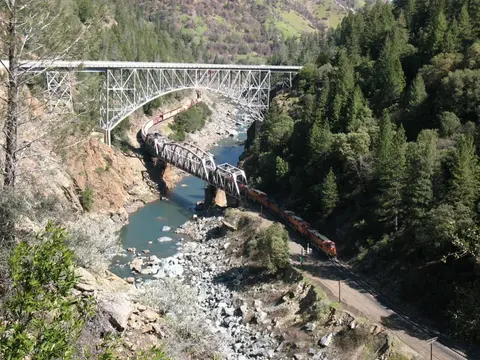 A train crosses a bridge spanning a rocky river gorge with a highway bridge above.