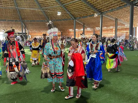People in traditional regalia gather inside a large covered pavilion with a turf floor.