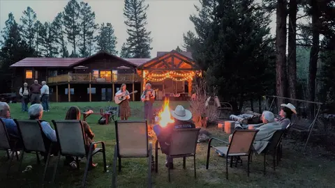 People sitting around a campfire listening to musicians play guitars outside a lodge at dusk.