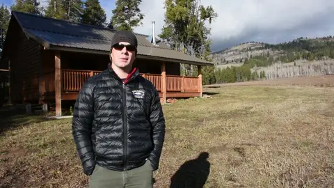 A man stands in a field in front of a rustic cabin with mountains in the background.