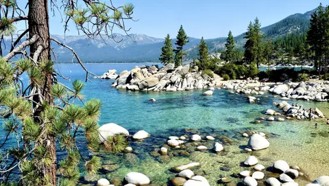 A scenic view of a clear blue lake with large boulders and pine trees along the shore.