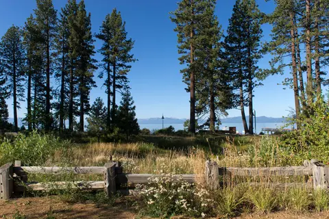 A scenic view of a lake and distant mountains framed by tall pine trees and a wooden fence.