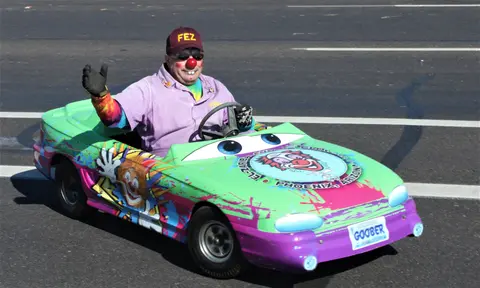 A clown waves while driving a brightly painted miniature car on a street.