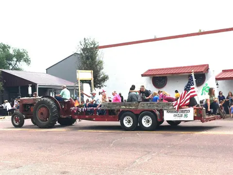 A tractor pulls a parade float carrying people and American flags.