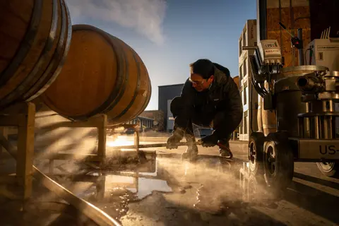A person works outdoors with steam and equipment near wooden barrels.