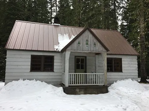 A cabin with snow on the ground and roof in a forest.