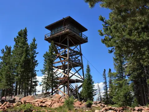 A tall wooden fire lookout tower stands among pine trees and rocks under a blue sky.