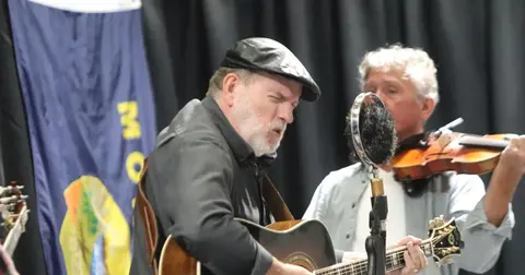 A man wearing a cap plays an acoustic guitar while another man plays the fiddle.