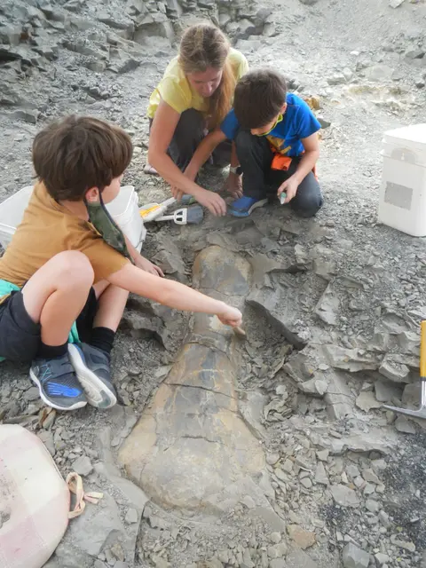 A woman and two children brush and excavate a fossil in the dirt.