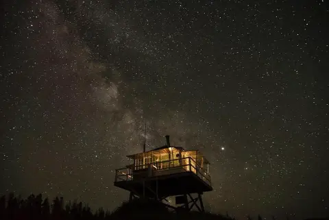 A fire lookout tower is lit up at night under a starry sky.