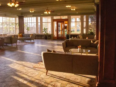 An indoor lobby or sitting room with sofas and large windows illuminated by sunlight.