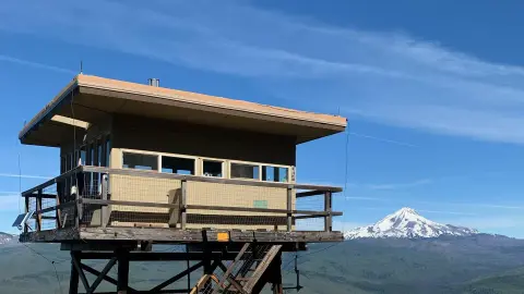 A lookout tower stands on a wooden platform with a snowy mountain in the background under a blue sky.