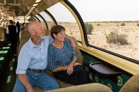 An older couple sits inside the dome car of a train looking out the window at a desert landscape.
