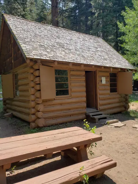 A wooden log cabin stands in a wooded area with a picnic table in the foreground.