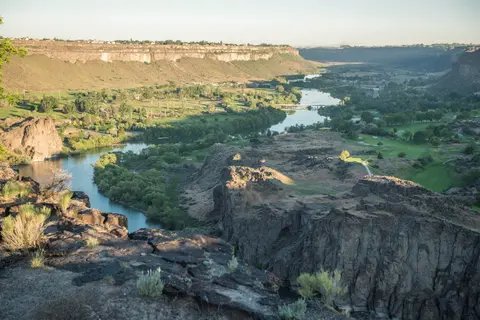 A wide canyon with a winding river running through it and green fields on its floor.