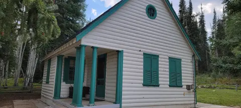 Exterior view of a small white log cabin with green trim and pillars in a wooded area.