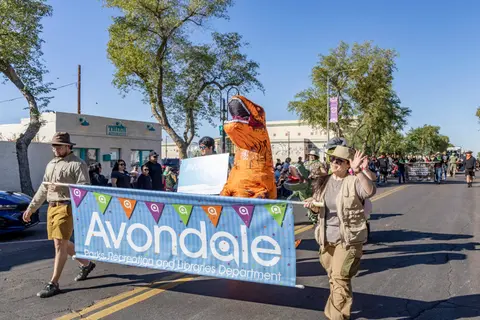 People in safari attire and an inflatable dinosaur carry a banner in a parade on a street.