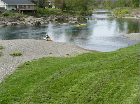 People kayaking on a river near a grassy bank and a house.