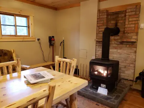 An interior view of a cabin featuring a dining table and a wood-burning stove against a brick hearth.
