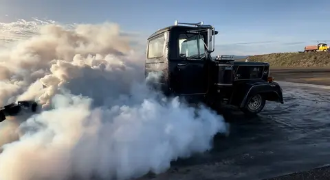 A dark truck doing a burnout on an asphalt surface, producing thick white smoke.