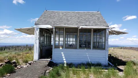 A small white building with many windows sits on a grassy landscape under a blue sky.