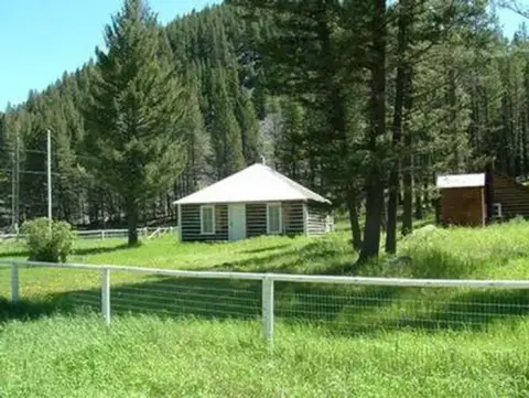 A cabin nestled in a grassy clearing surrounded by trees and mountains.