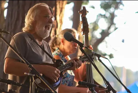 A man plays a mandolin and sings into a microphone while a woman plays bass behind him.