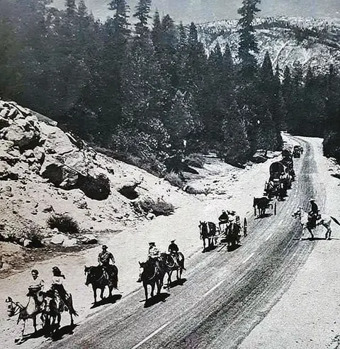 A historic black and white photo shows a line of horses, riders, and carriages traveling along a mountain road.