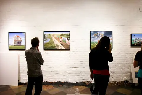 People view framed paintings on a white brick wall inside a gallery.
