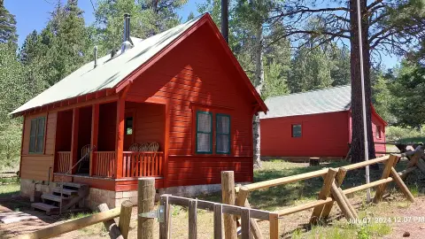 A red cabin with a porch stands in a wooded area behind a wooden fence.