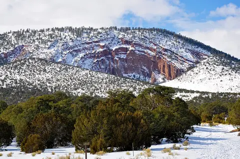 A snow-covered landscape featuring red rock cliffs and sparse trees under a partly cloudy sky.