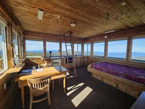 The interior of a wooden cabin features a bed, a table, chairs, and windows offering a scenic view.