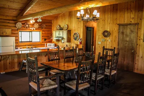 An interior view of a rustic log cabin kitchen and dining area with a long wooden table and chairs.