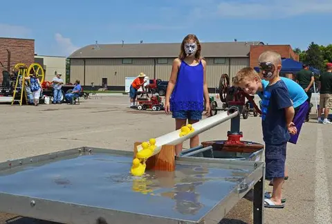 Children play with a water pump and toy ducks in a large outdoor lot.