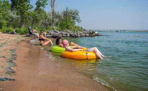 People relax on inner tubes in the water near a sandy beach.