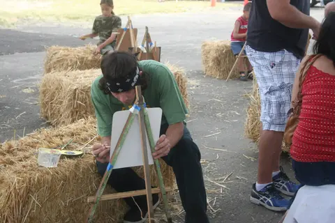 People sit on hay bales outdoors and paint on small easels.
