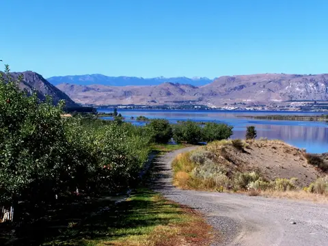A dirt road winds past orchards toward a lake and distant mountains under a clear blue sky.