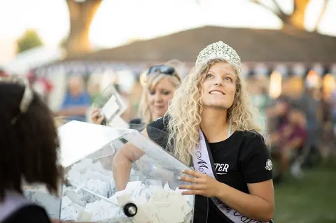 A woman wearing a tiara and sash reaches into a raffle drum.