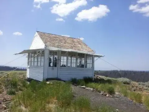 A small white building with many windows sits on a grassy hill under a blue sky.