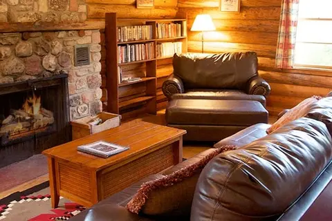 A cozy rustic living room features a stone fireplace, leather furniture, and a wooden bookshelf.
