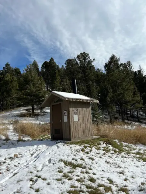 An outdoor toilet building sits in a snowy, wooded landscape under a cloudy sky.