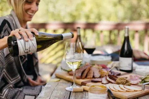 A smiling woman pours wine into a glass at an outdoor table set with food.