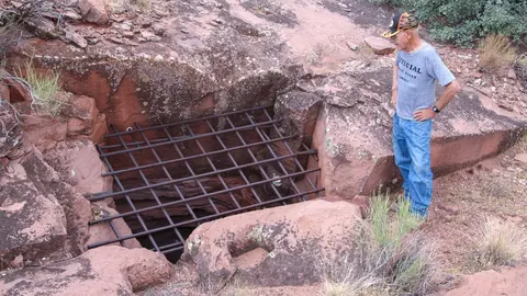 A man stands next to a metal grate covering a deep opening in the rocky ground.