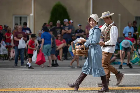 Two people in historical costumes walk along a street in front of a crowd of onlookers.