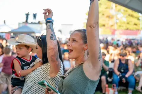 People cheer with their arms raised at an outdoor event.